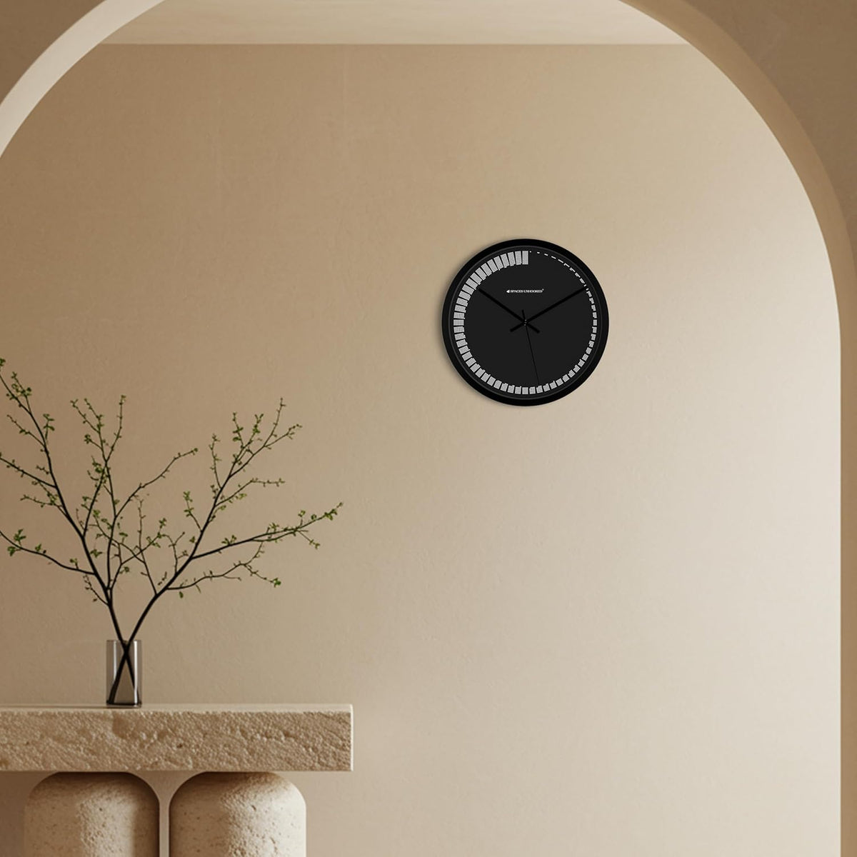 Black wall clock on a beige wall with a decorative plant on a stone ledge.