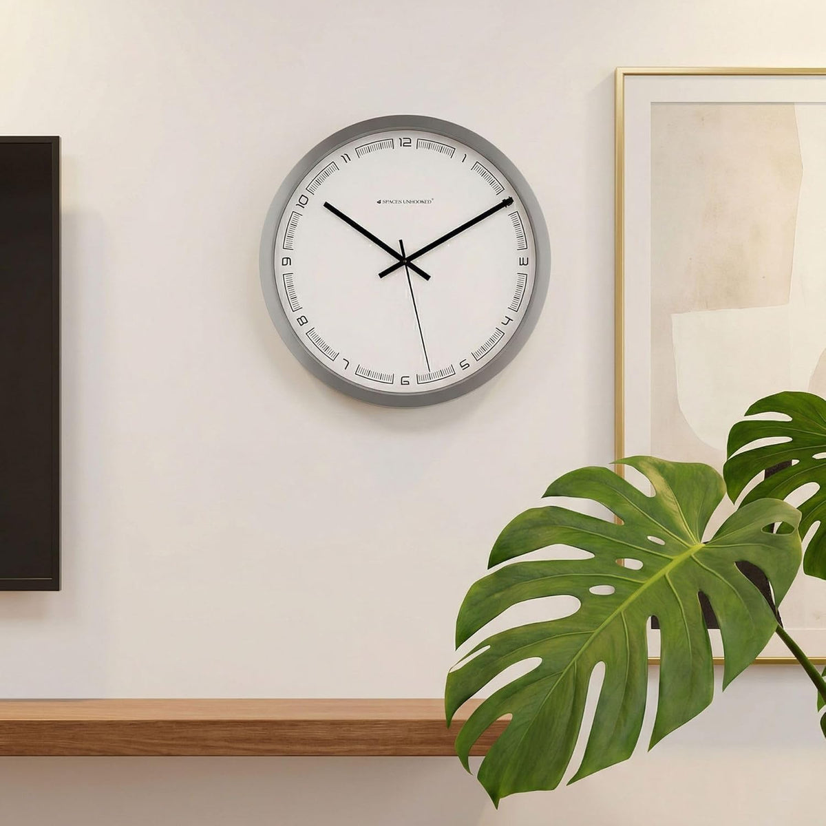 Wall clock on a wall with a plant and framed picture in the background