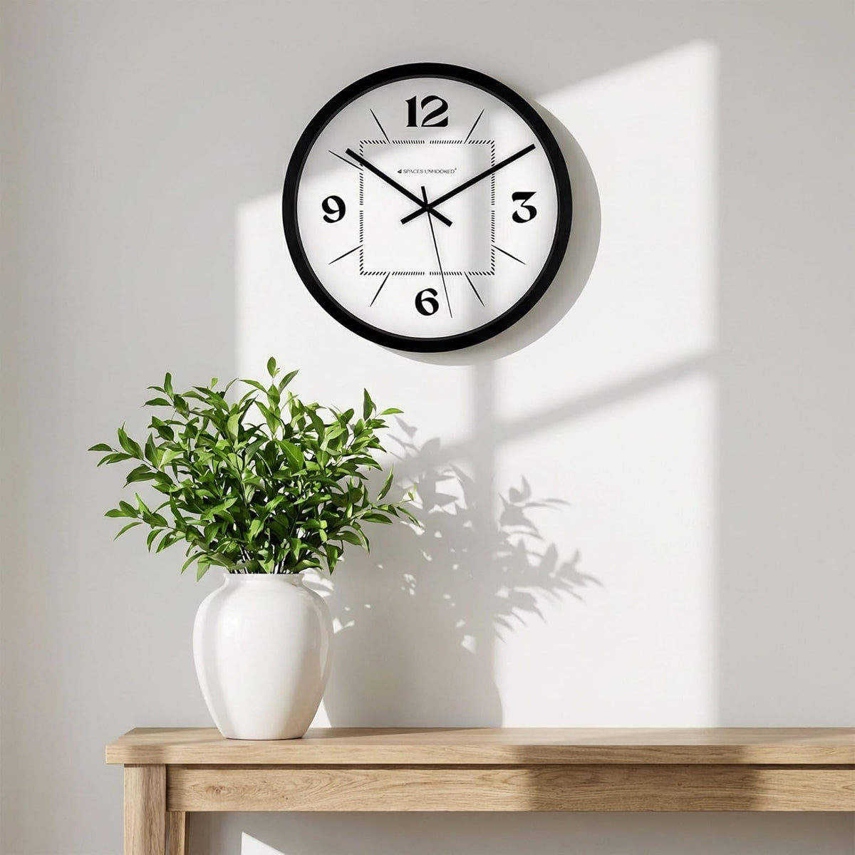 Wall clock on a white wall with a plant on a wooden surface below.