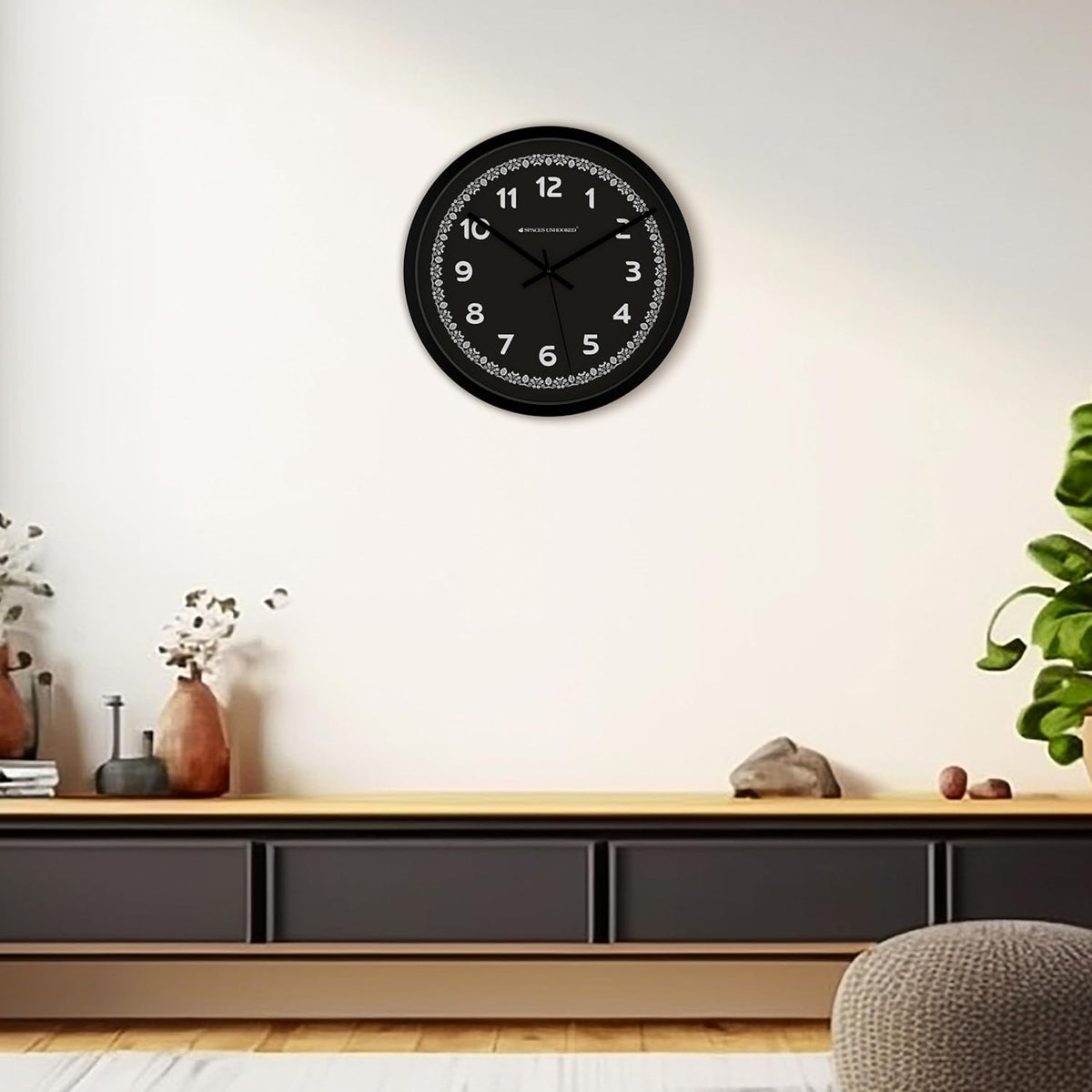Black wall clock on a white wall above a wooden shelf with decorative items.