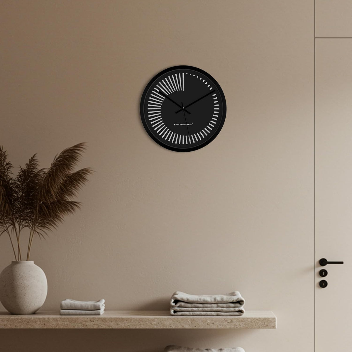 Black wall clock on a beige wall with a shelf below featuring folded towels and a vase.
