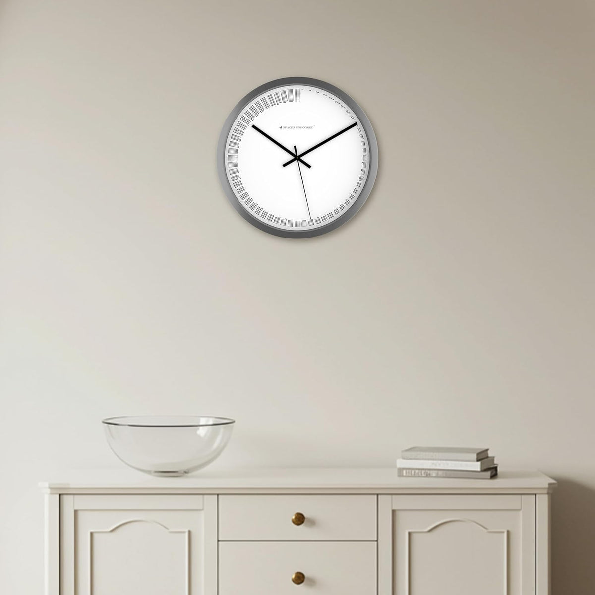Wall clock on a plain wall above a white cabinet with a glass bowl and books.