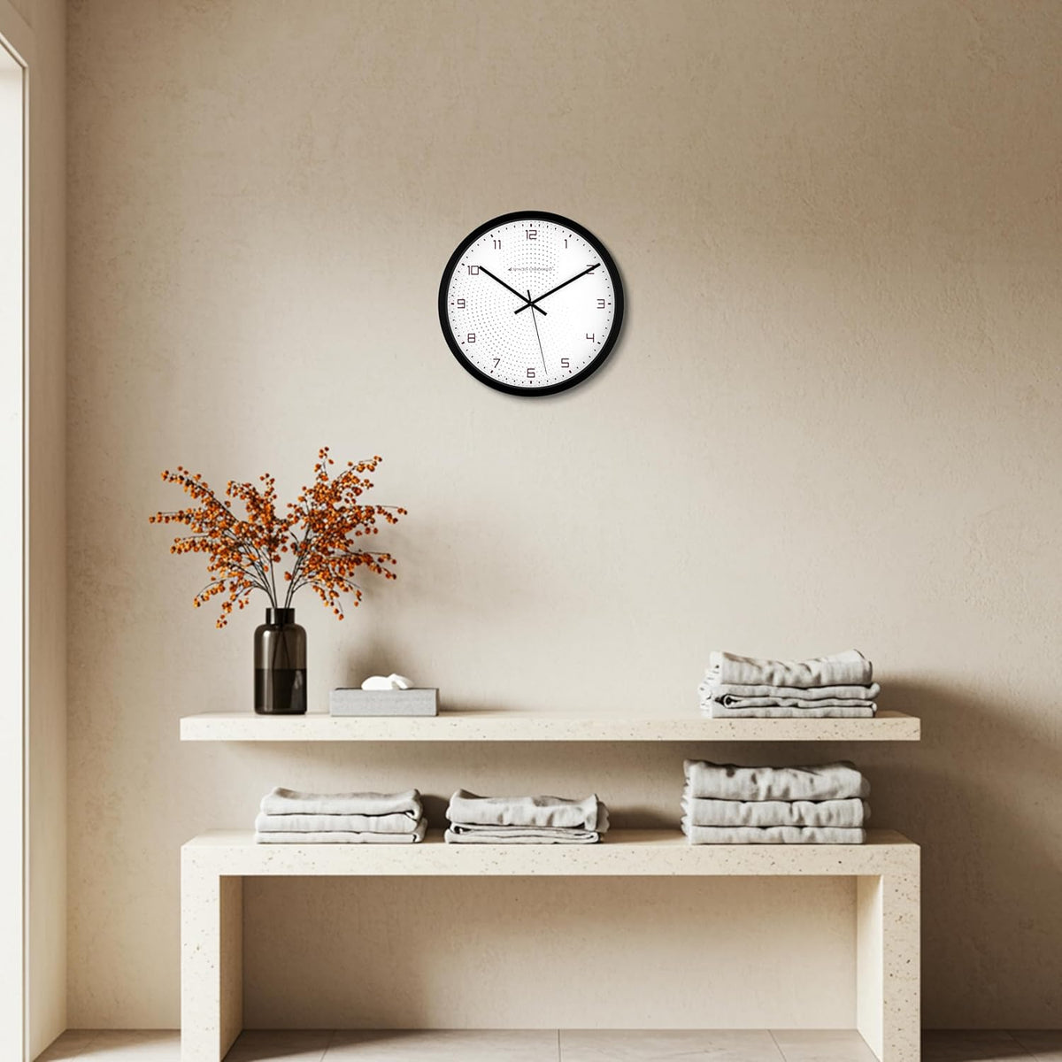 Wall clock on a beige wall above a shelf with folded towels and a vase of flowers.