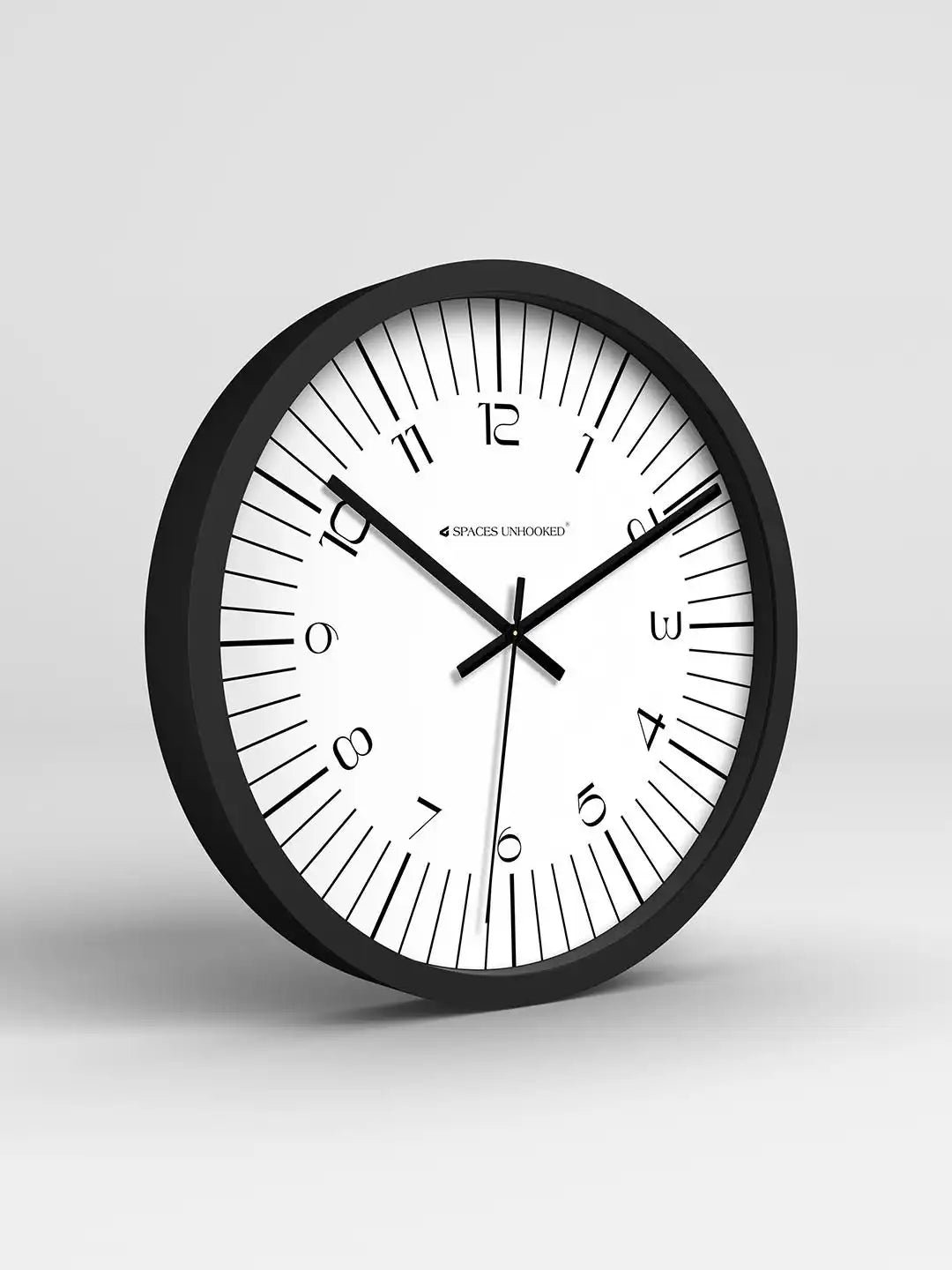 Black-framed clock with bold tick marks displaying 10:11, leaning on a white table beside a book, wooden ornament, and potted plants.