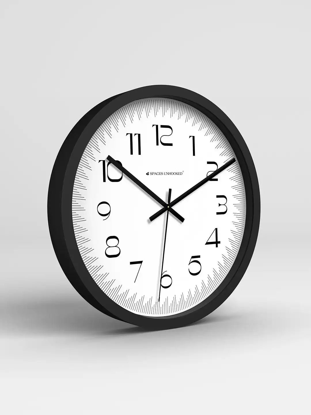 Close-up of a black-framed wall clock with stylized numerals showing 10:11, set on a wooden surface with green leaves blurred in the foreground.