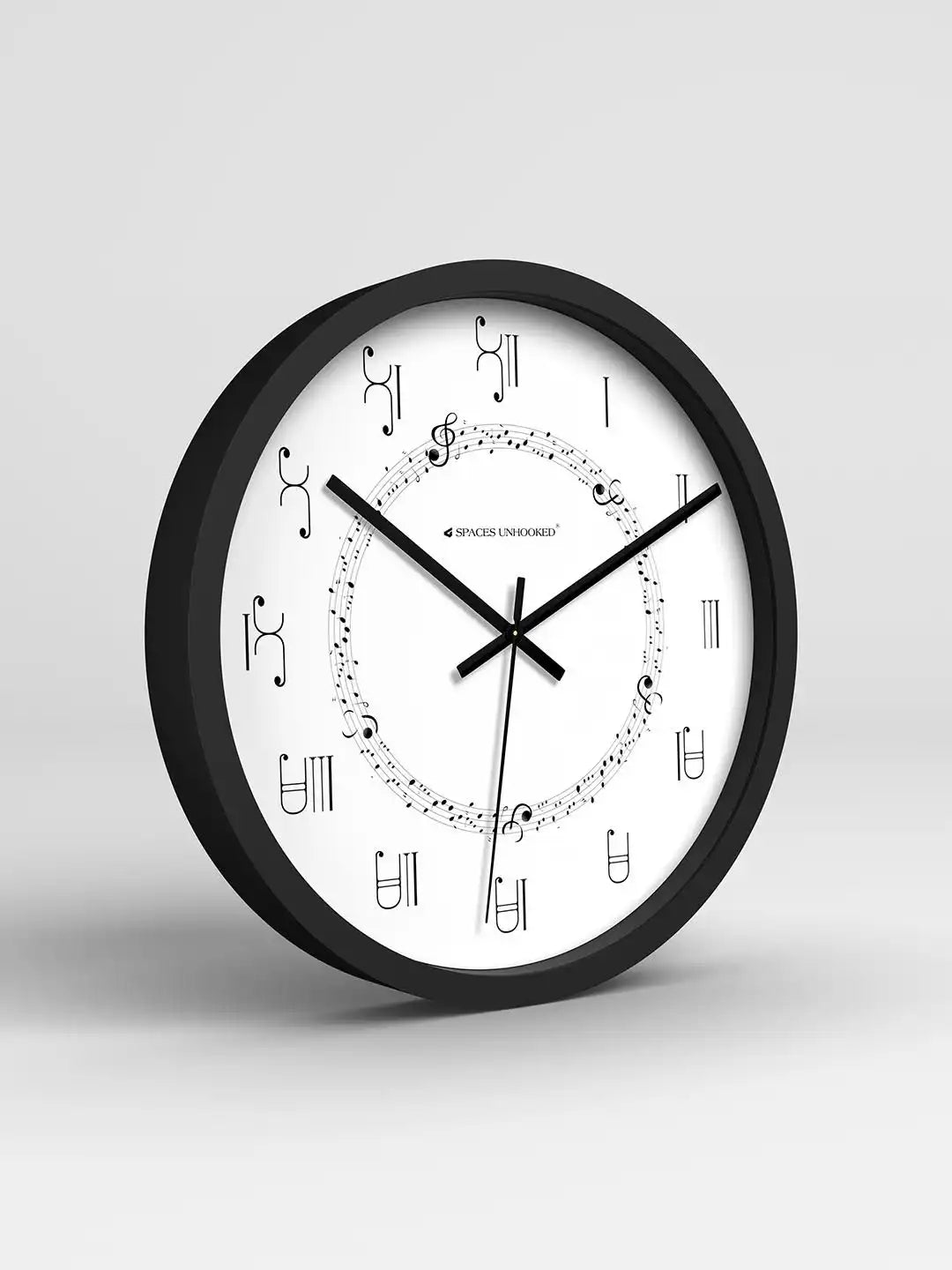 Black-framed clock with music-note ring design and Roman numerals showing 10:11, on a wooden table beside a small potted plant against sheer white curtains.