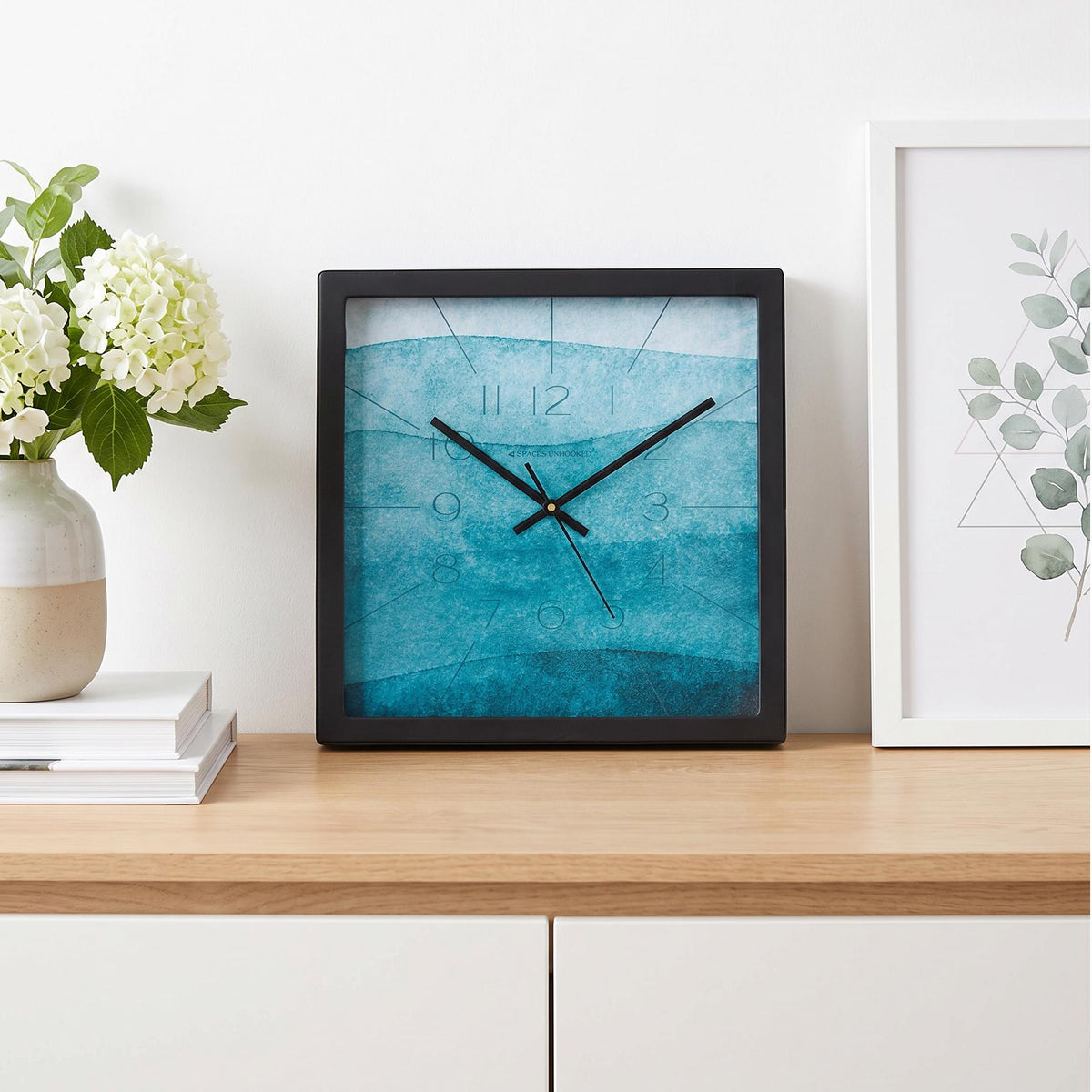 Black square frame and clear cover of a teal watercolour wall clock, close-up view
