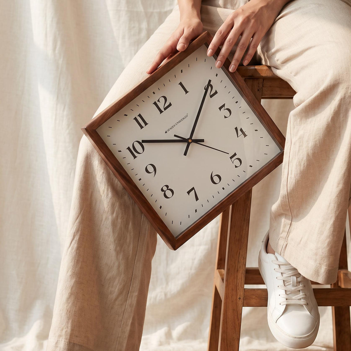 Square wooden wall clock with classic numerals hanging above a sofa in a bright living room