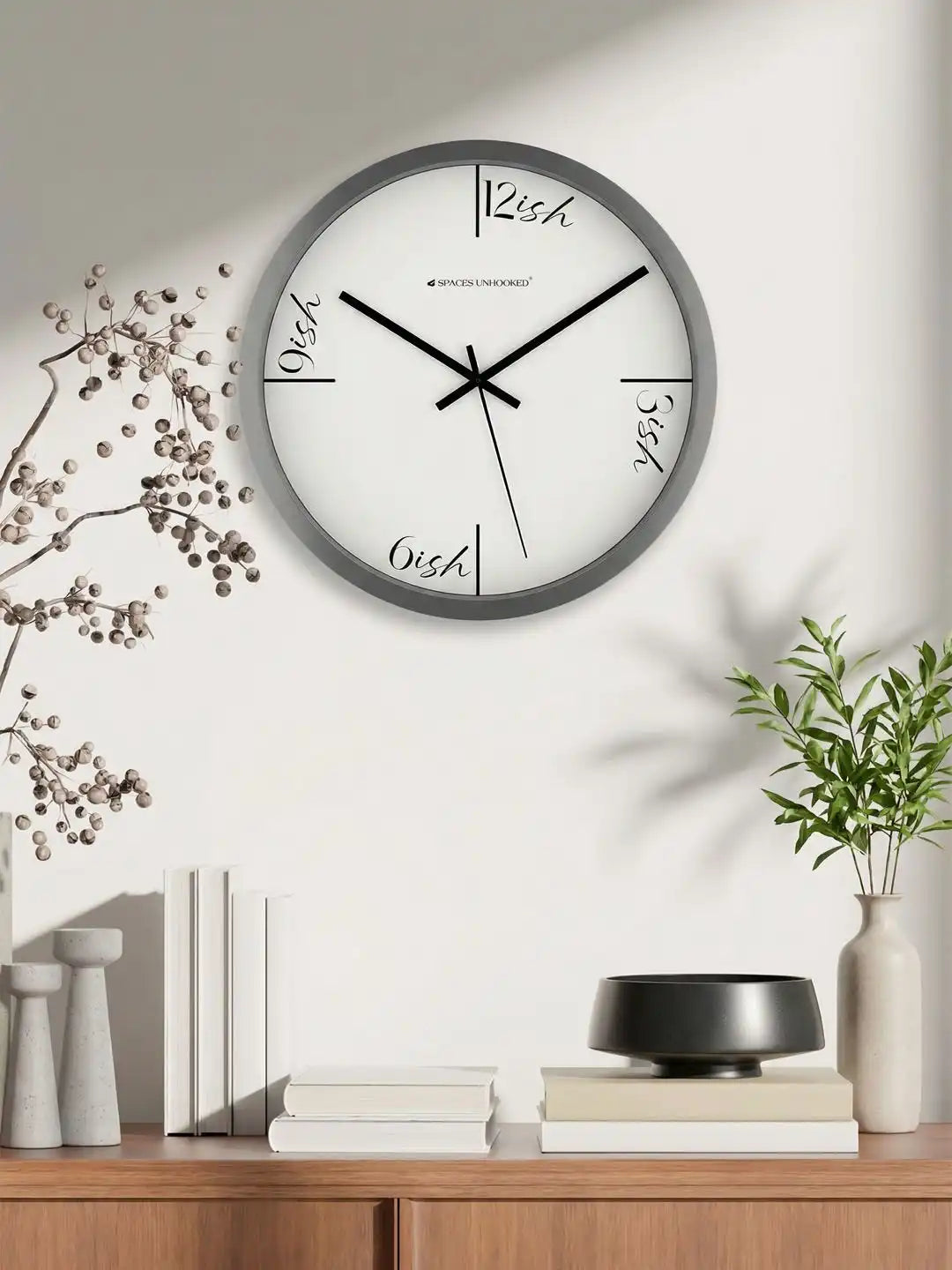 Grey wall clock with "ish" dial design mounted above a wooden sideboard with dried branches, books, and a vase of greenery