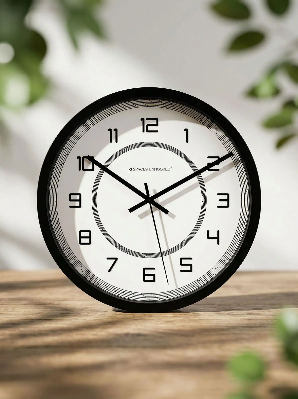 Black wall clock with white dial displayed on a wooden table with green plant leaves in the background
