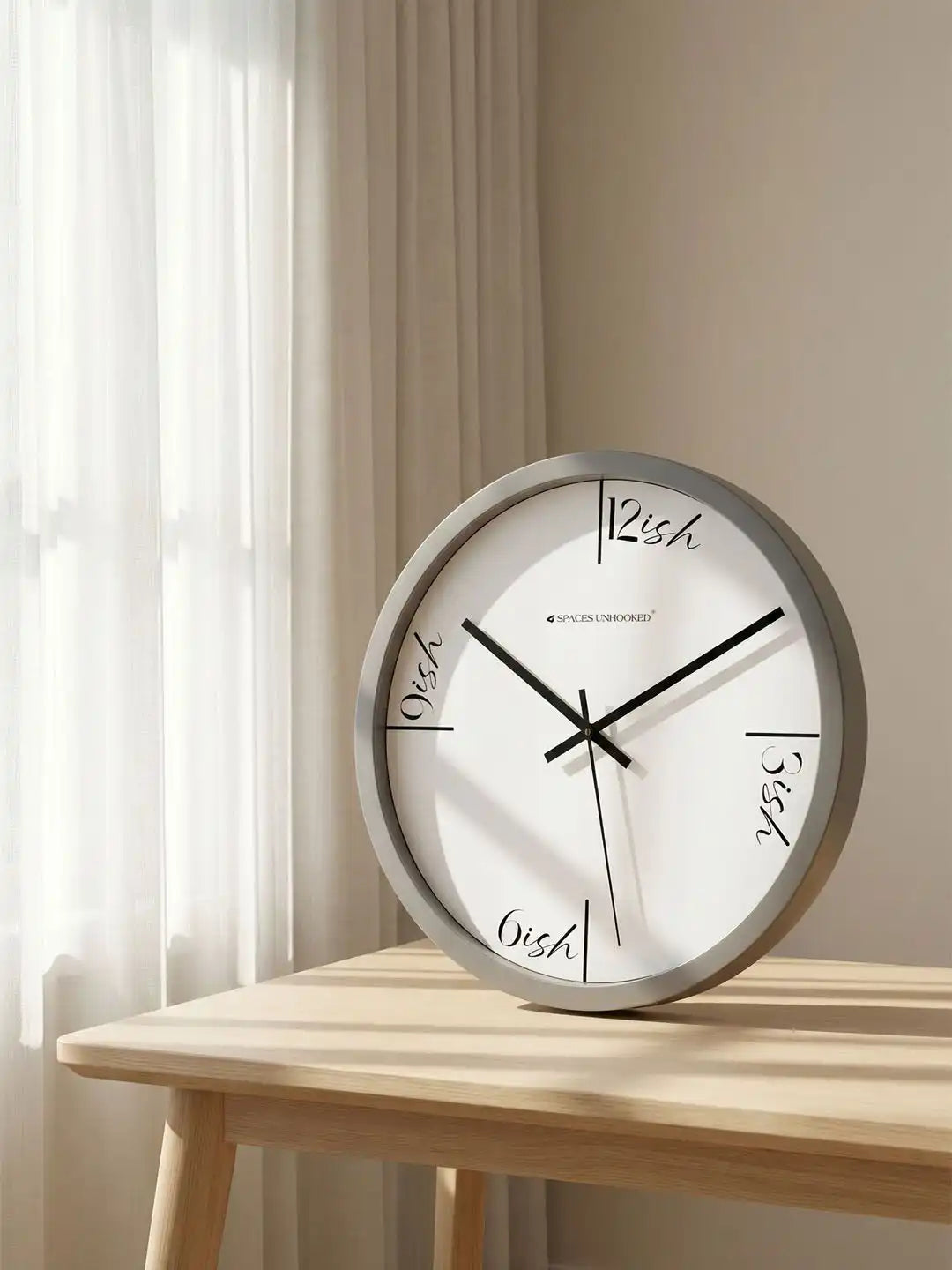 Grey wall clock with "ish" dial design on a beige wall beside sheer curtains, resting on a light wood table