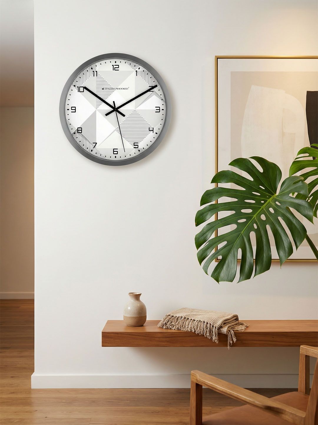 Wall clock on a white wall with a plant and wooden shelf in the background